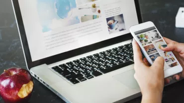 Persona usando un teléfono y una laptop en una mesa con una manzana y suculenta.