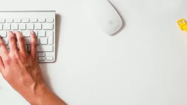 Manos escribiendo en un teclado junto a un reloj amarillo.