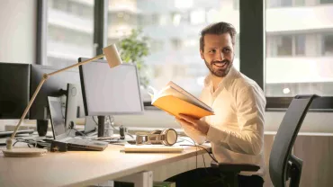 Persona en una oficina sosteniendo un libro, rodeada de computadoras.