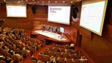 Personas asistiendo a una charla empresarial en un auditorio con panelistas en el escenario.