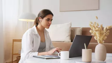 Persona trabajando en laptop desde casa, mesa con cuaderno y taza.