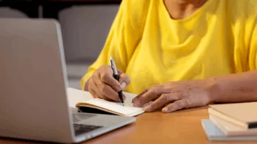 Persona escribiendo en un cuaderno junto a una laptop.