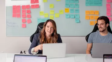 Personas trabajando con computadoras en una oficina con una pizarra llena de notas adhesivas.