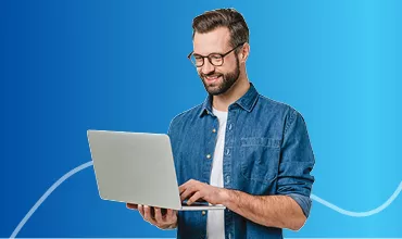 Hombre con gafas sonriendo, de pie, sosteniendo un portátil en alto. Parece feliz y entusiasmado.